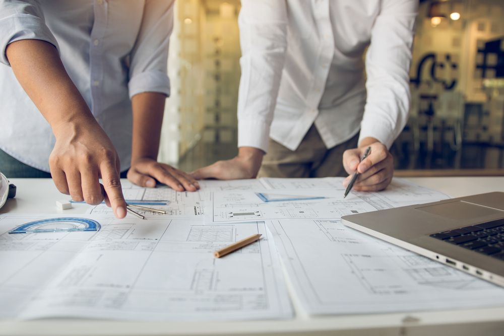 a close up of two males pointing to project designs on a table with pencils and triangular set squares and protractor