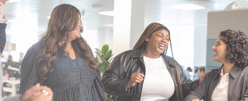 Three women in an office setting are smiling and laughing together, suggesting a positive and inclusive work environment. They appear to be having a friendly and joyful conversation, promoting a sense of camaraderie and diversity.