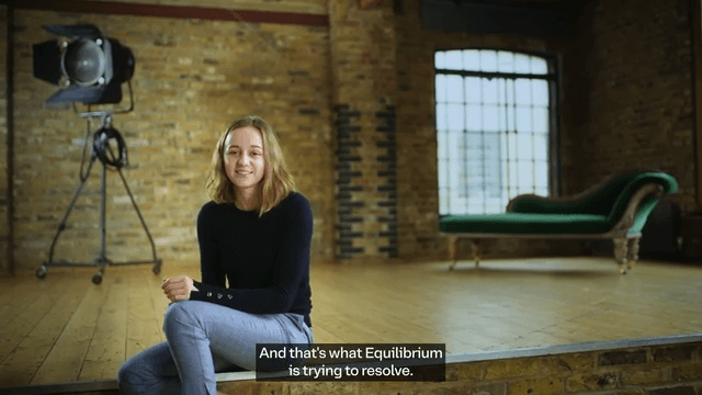 A woman sits on a wooden stage in a loft-style room with brick walls and large windows. Behind her is a vintage green chaise lounge and a studio light. The subtitle reads, "And that's what Equilibrium is trying to resolve.