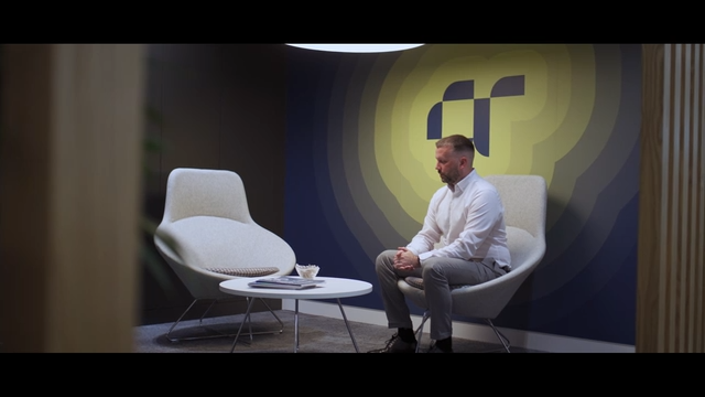 A man in a white shirt sits in a modern office lounge area with minimalist chairs and a round table. The wall behind him features a large abstract logo with concentric gradient circles in yellow and blue tones.