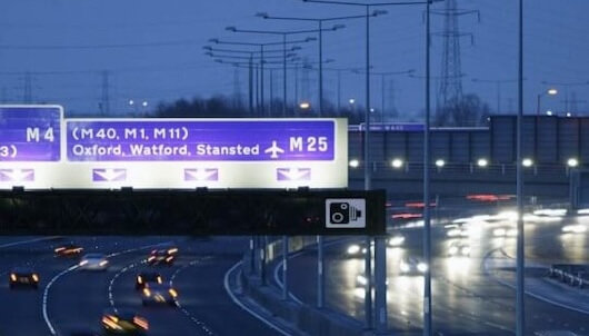 A nighttime view of a busy highway with multiple lanes of traffic and a large overhead road sign indicating directions to the M4, M40, M1, M11, Oxford, Watford, Stansted, and M25. Headlights and taillights of vehicles create light trails.