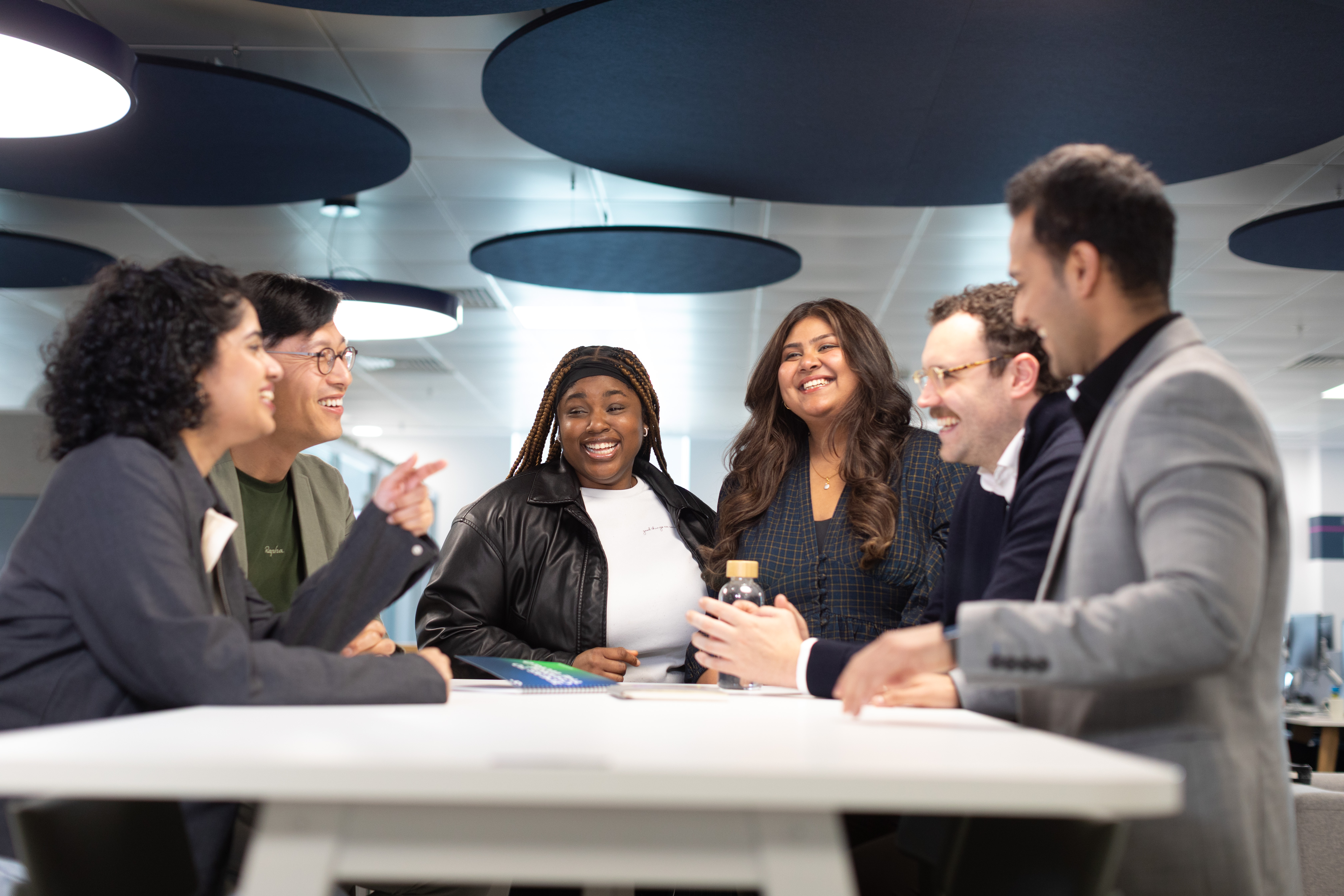 Diverse team gathered around a table in a collaborative meeting, all smiling and engaged in conversation in a contemporary workspace.