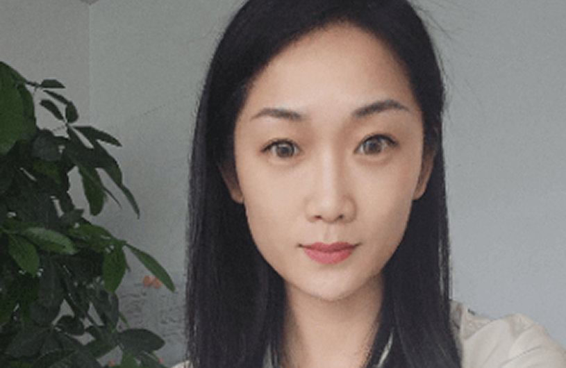 Portrait of an Asian woman with long black hair, wearing a light top, standing indoors with natural lighting and a green plant in the background.
