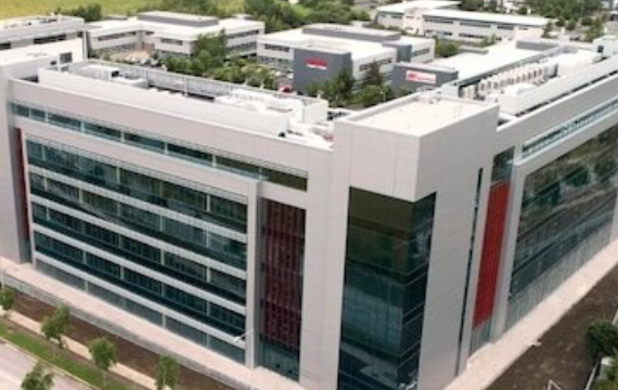 Aerial view of a modern office building in a business park surrounded by greenery and roads.