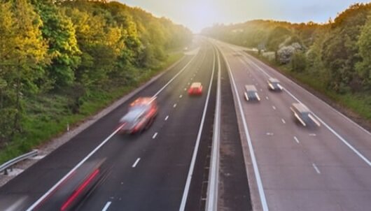 A highway with multiple lanes surrounded by green trees on both sides, with cars moving at speed, captured with motion blur during sunset.
