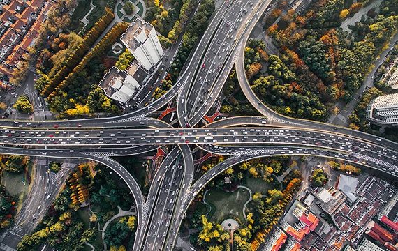 Aerial view of a large, complex highway interchange surrounded by trees and buildings, with multiple lanes of traffic moving in different directions.