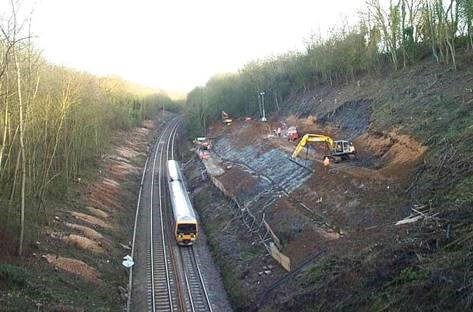 A train travels along a railway line through a cutting, while an excavator and workers perform slope stabilization or construction work on the hillside beside the tracks. Trees line both sides of the scene.