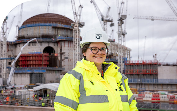 Woman in a high-visibility safety jacket and hard hat smiling at a construction site with cranes and a large industrial structure in the background.