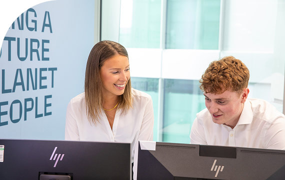 Two young professionals collaborating and smiling at their computer screens in an office environment.