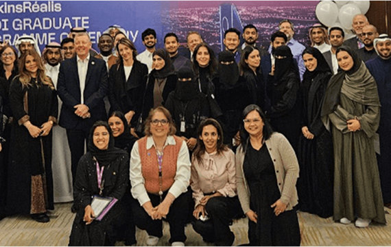 Group photo of diverse men and women at the AtkinsRéalis Saudi Graduate Programme Ceremony, standing in front of a backdrop featuring a cityscape with the text "Saudi Graduate Programme Ceremony." The group includes individuals in business attire, traditional Saudi dress, and hijabs, gathered in a formal indoor setting.