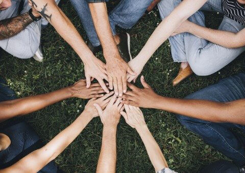 A group of diverse people sitting in a circle on grass, placing their hands together in the center, symbolizing unity and teamwork.
