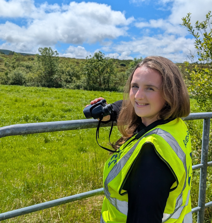 Caroline standing at a metal gate in a field, looking over her shoulder and smiling