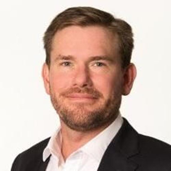 A man with short hair and a trimmed beard, wearing a suit jacket and white shirt, posed against a white background.
