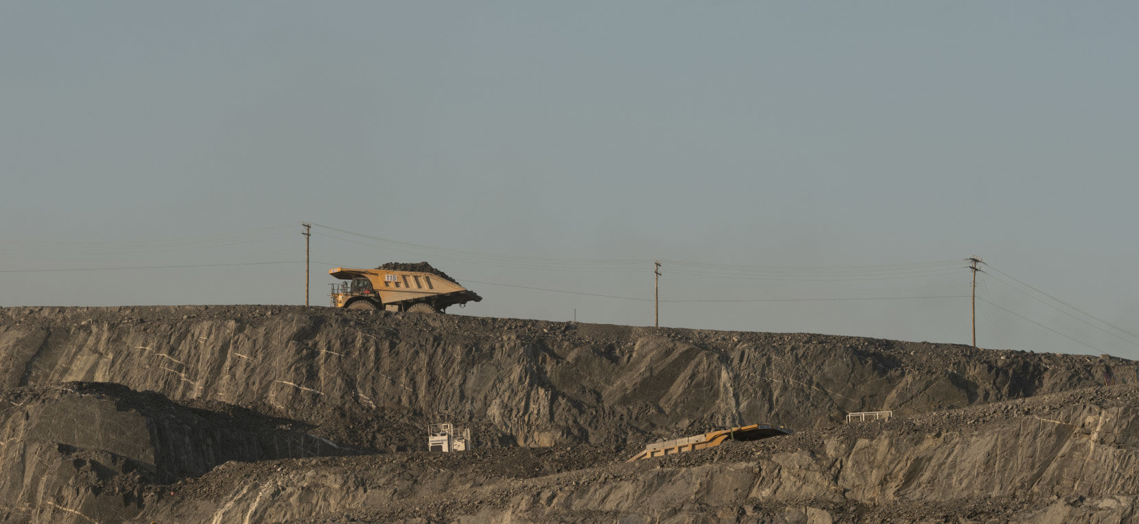 Large mining dump trucks driving along the edge of an open-pit mine with rocky terrain and power lines in the background.