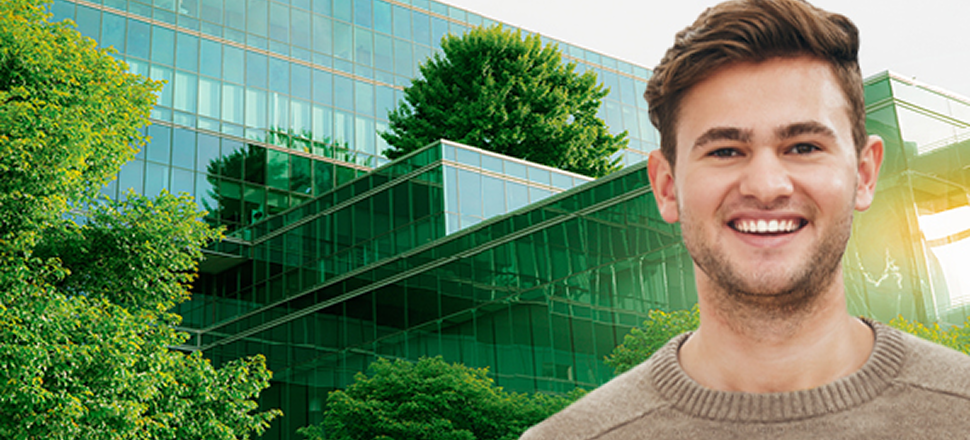 Smiling young man standing in front of an aerial view of a large, green highway interchange with a city skyline in the background.