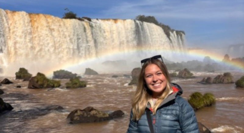 A smiling picture of Maisie standing in front of a water falls and a rainbow falling in between