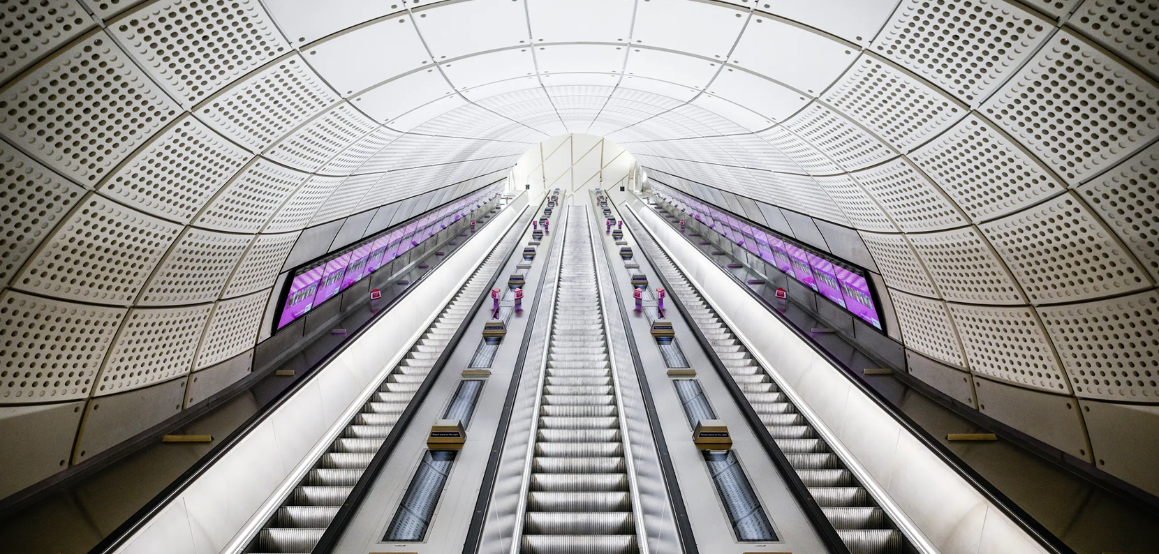 Modern underground escalators leading upward inside a futuristic, curved metallic tunnel.