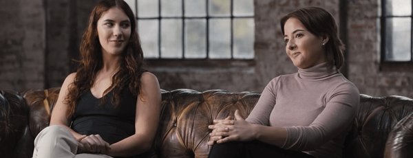 Two women sitting on a leather couch in a casual conversation, with large industrial windows in the background.