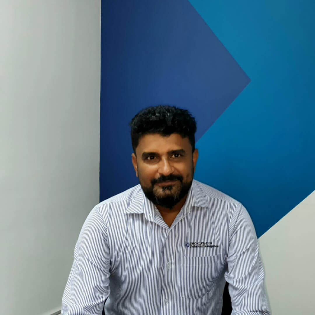 The image shows a man with short curly hair and a beard, sitting indoors and wearing a light striped button-up shirt with a small logo. He’s looking at the camera with a slight smile.