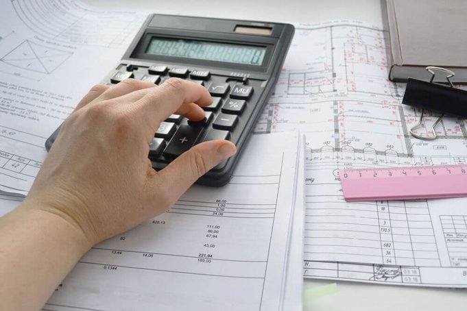 Hand using a calculator on top of architectural drawings, papers, and a pink ruler.