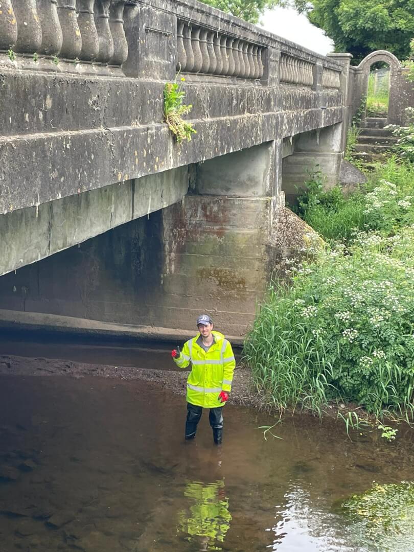 Person in high-visibility gear standing in shallow water under a stone bridge.