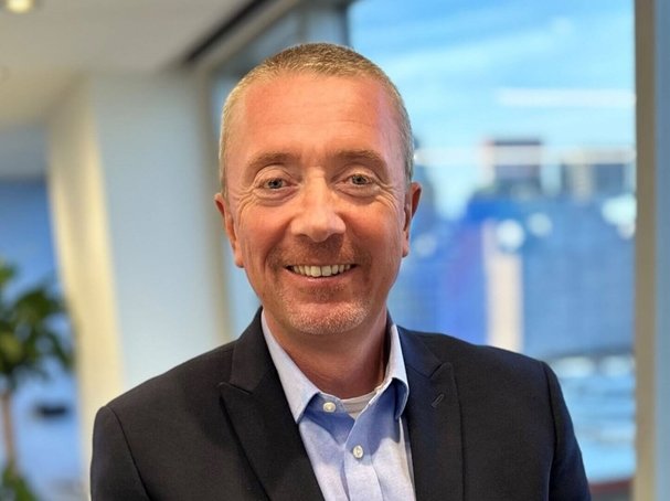 Smiling man in a blazer and blue shirt standing indoors with cityscape background.