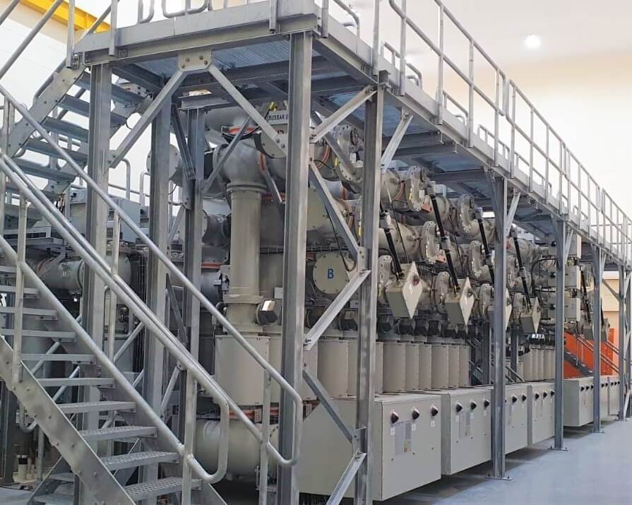 Large industrial gas-insulated switchgear (GIS) installation inside a facility, featuring multiple high-voltage components, control panels, and interconnected pipes mounted on a metal platform with access stairs and walkways.