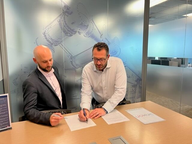 Two men in business attire sitting at a conference table reviewing and signing documents in a modern office with frosted glass walls and technical drawings in the background.