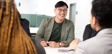 man wearing green jacket smiling broadly at a colleague over a desk