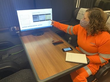 Alex on a train, wearing orange protective gear and pointing at a laptop screen