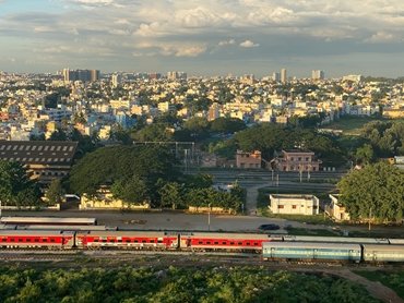landscape image of Bengaluru including city buildings, green spaces and trains on tracks in the foreground