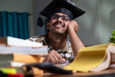 A young graduate wearing a graduation gap and glasses, sat in front of a pile of books.