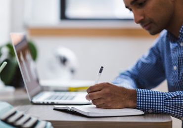man in blue shirt writing on a notepad next to an open laptop