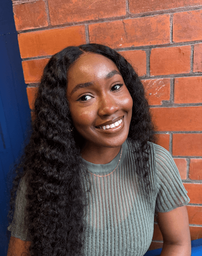 Smiling woman with long curly hair sitting in front of a red brick wall.
