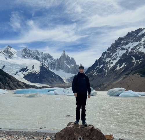 A picture of Tom standing on a rock, in front of ice caped mountains and a water stream of ice blocks