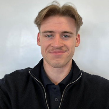 Young man with light brown hair, styled neatly, wearing a dark zip-up sweater over a navy polo shirt, smiling slightly in front of a plain white background.